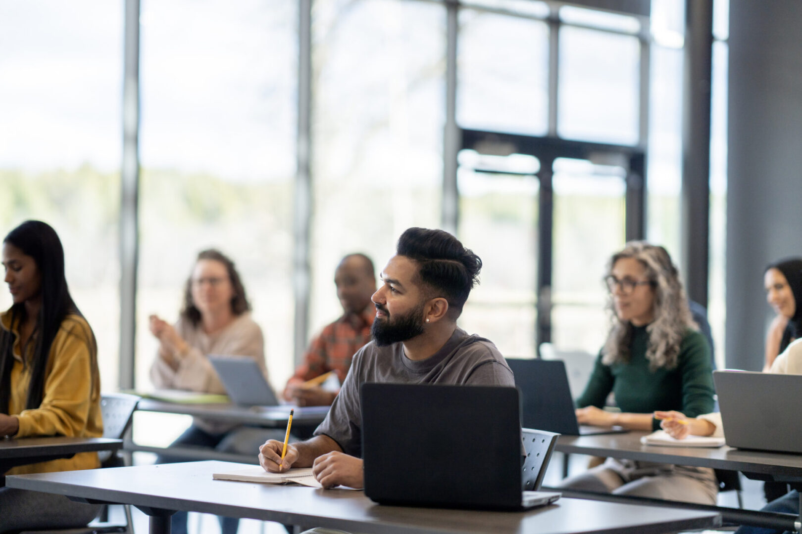 A large group of mature adults are seen sitting at desk with wither laptops and books open as they study during a continuing education class.  They are each dressed casually and are listening attentively.