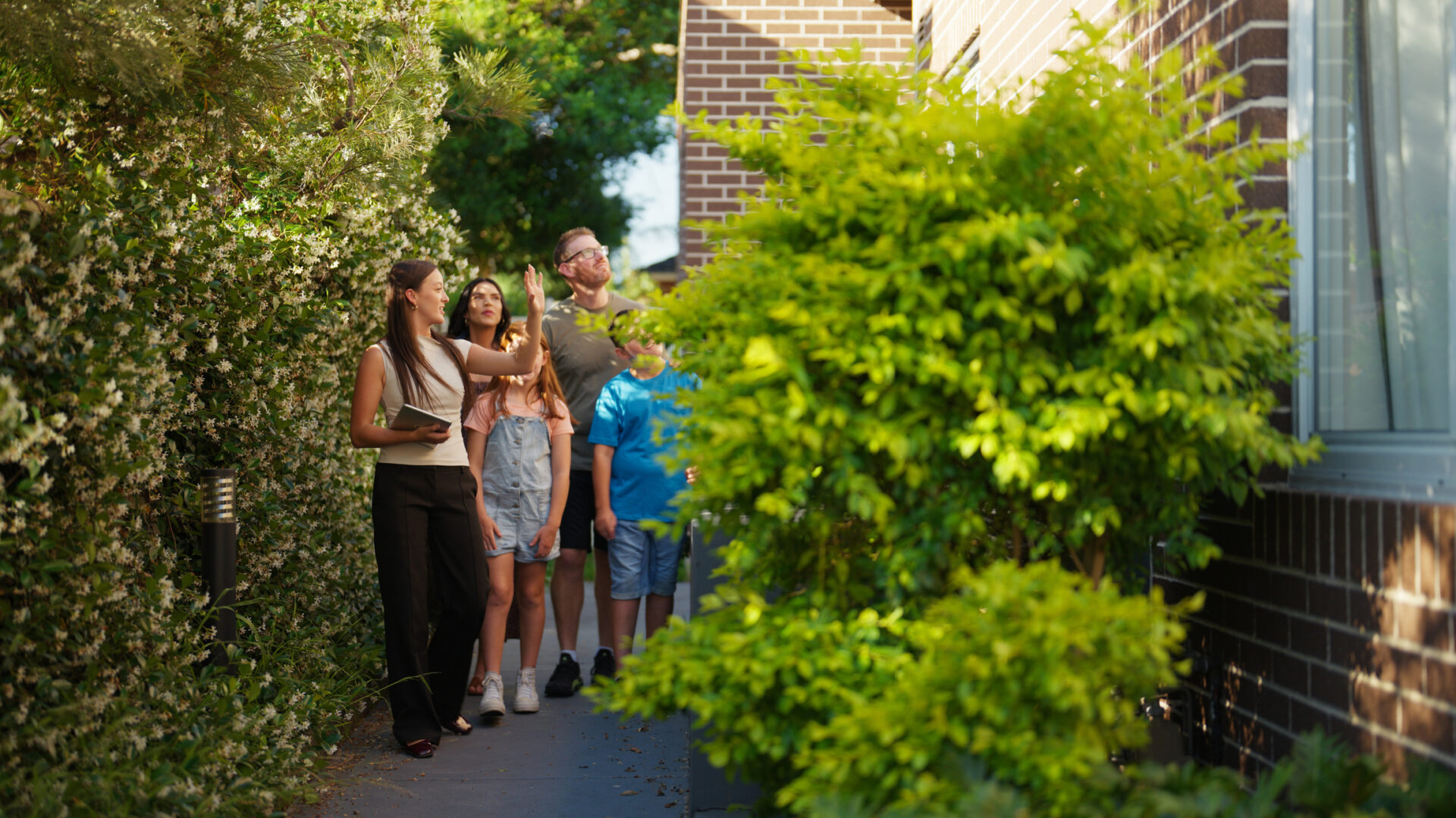 A real estate agent leads a family down a lush, green garden pathway alongside a modern brick home, showcasing the property’s serene outdoor features.