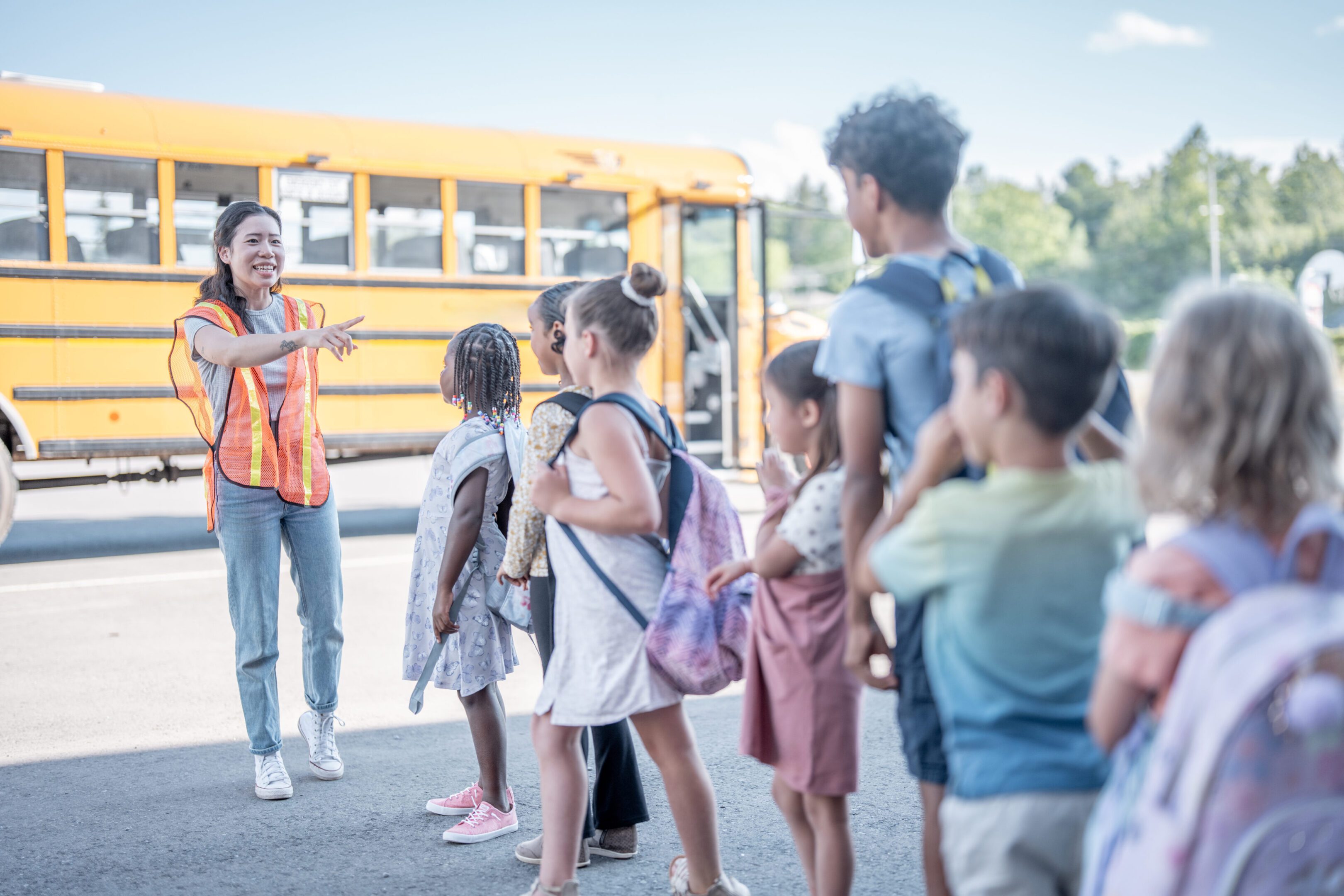 Children in a school setting engage with a supervising adult near a bright yellow school bus. The scene captures the essence of community, learning, and caring within an academic environment.