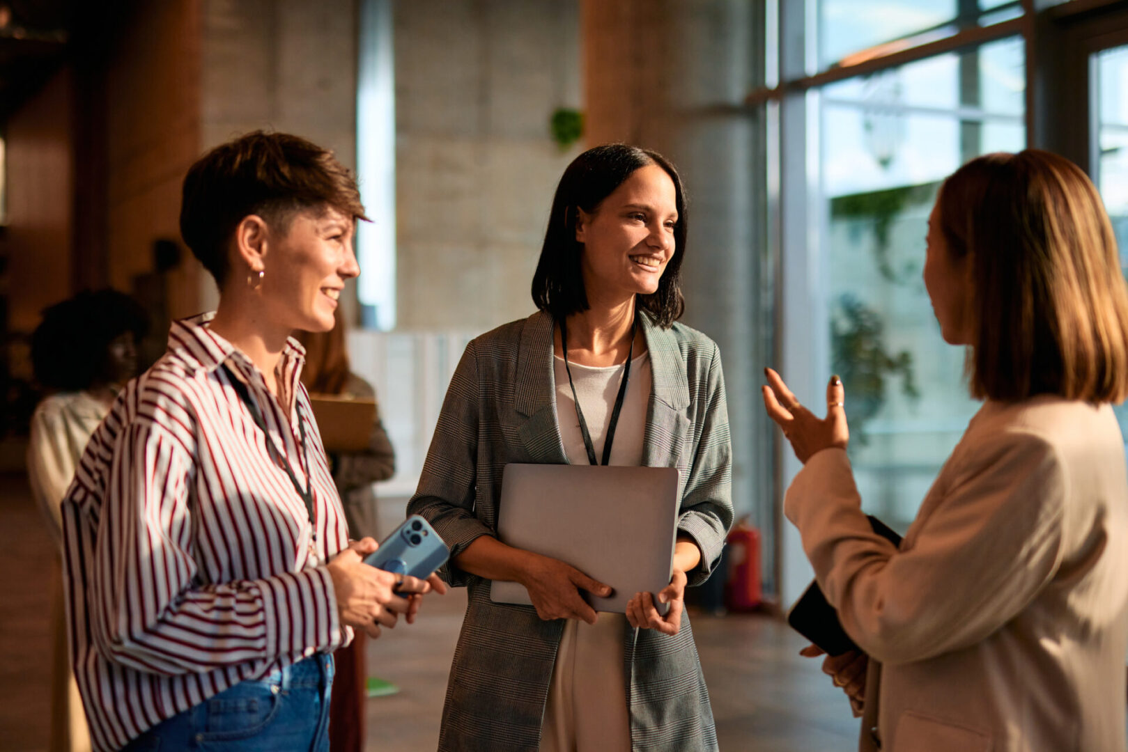 Three businesswomen standing in the lobby, engaging with a laptop, smartphone, and tablet, networking and discussing ideas during a conference break, fostering collaboration and connection