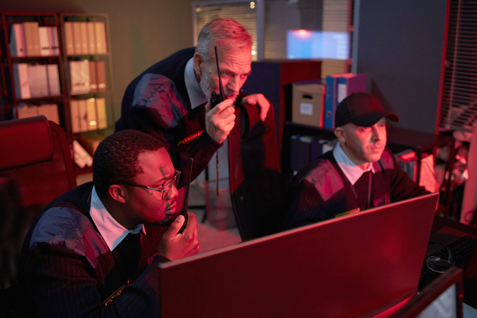 Middle aged Black man, Caucasian man, and middle aged man monitoring security footage on computer screens, communicating with walkie talkies in control room