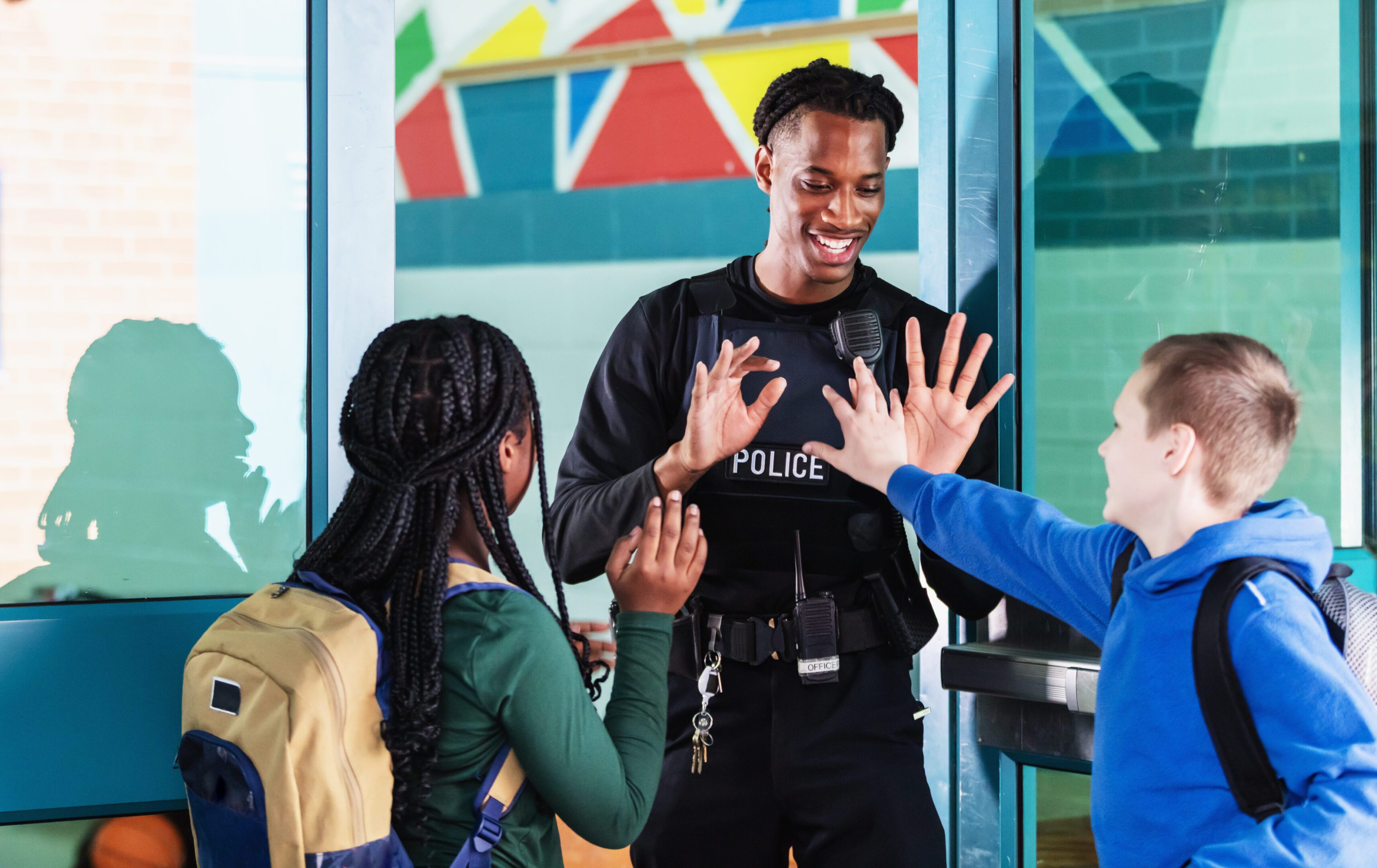 A school resource officer greets students arriving with their backpacks. They are standing face to face with the view over students' shoulders. The officer, a young African-American man, is giving a high-fives to the 10 year old girl and 12 year old boy.