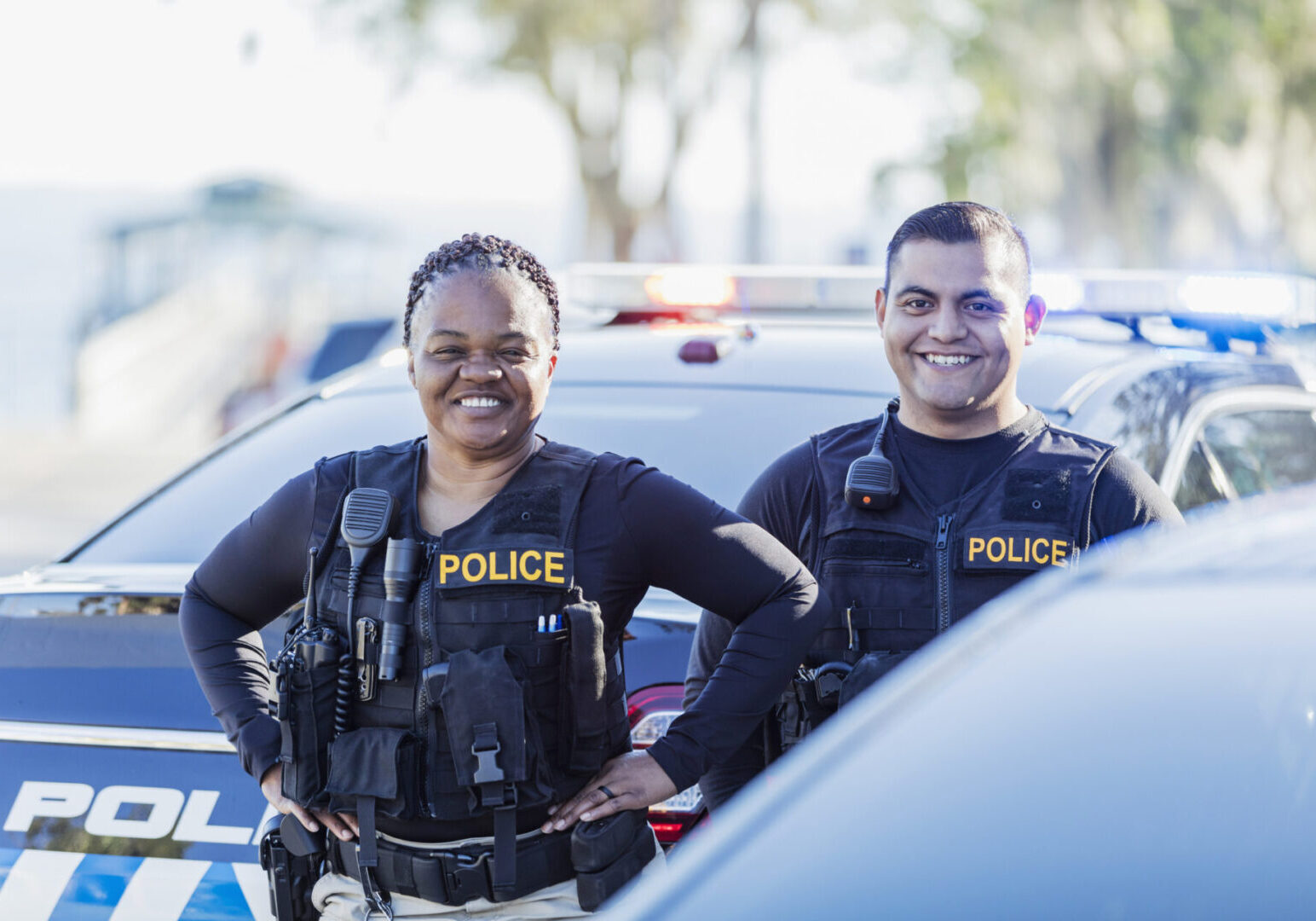 Two multi-ethnic police officers standing together next to their patrol car. The African-American policewoman is in her 40s and her partner is a young hispanic man in his 20s. They are smiling at the camera.