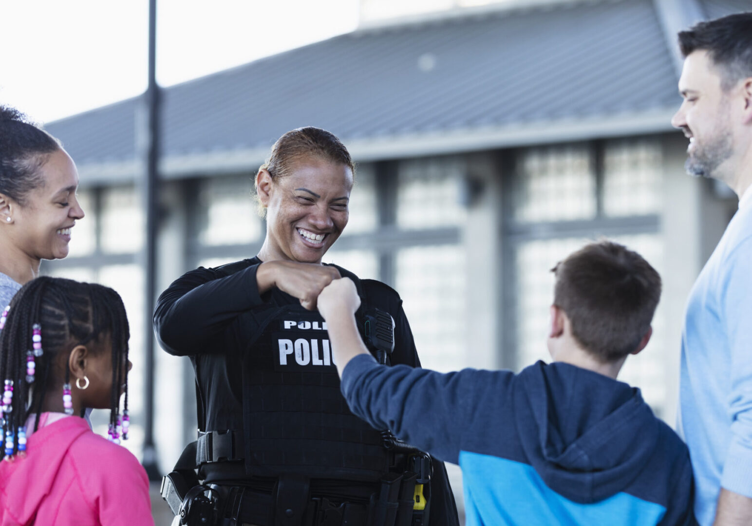 A multiracial police woman talking to a group of parents and children outdoors. standing outside a building. She is smiling at an 11 year old boy and they are bumping fists. The view is from over the boy's shoulder.