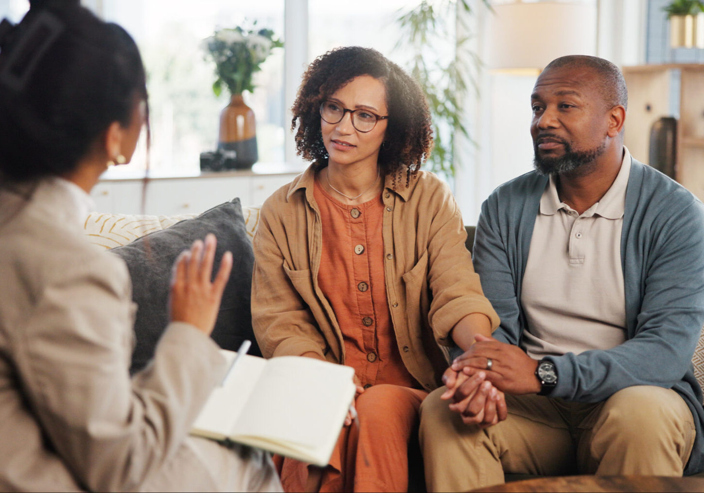 Couple, therapy and holding hands with discussion on sofa for guidance, communication and advice. Consultation, service and therapist with book in office for marriage counseling, support and progress