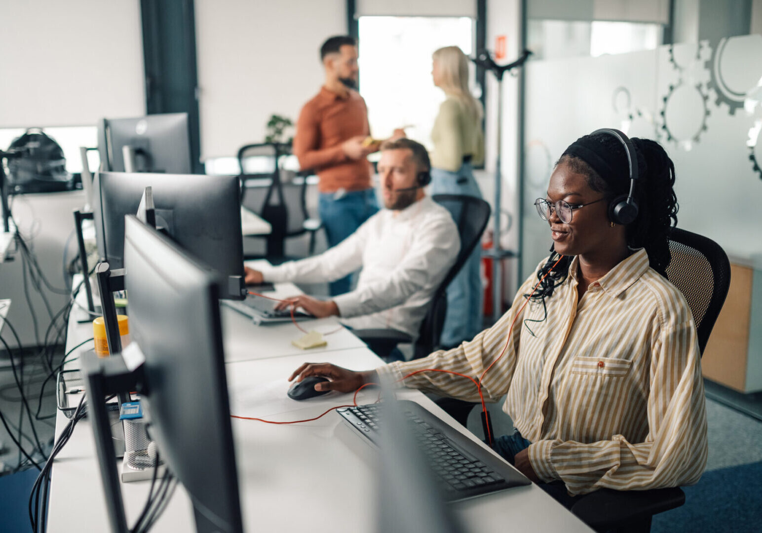 Black female support agent wearing headset is using computer while working in a call center with her colleagues in the background
