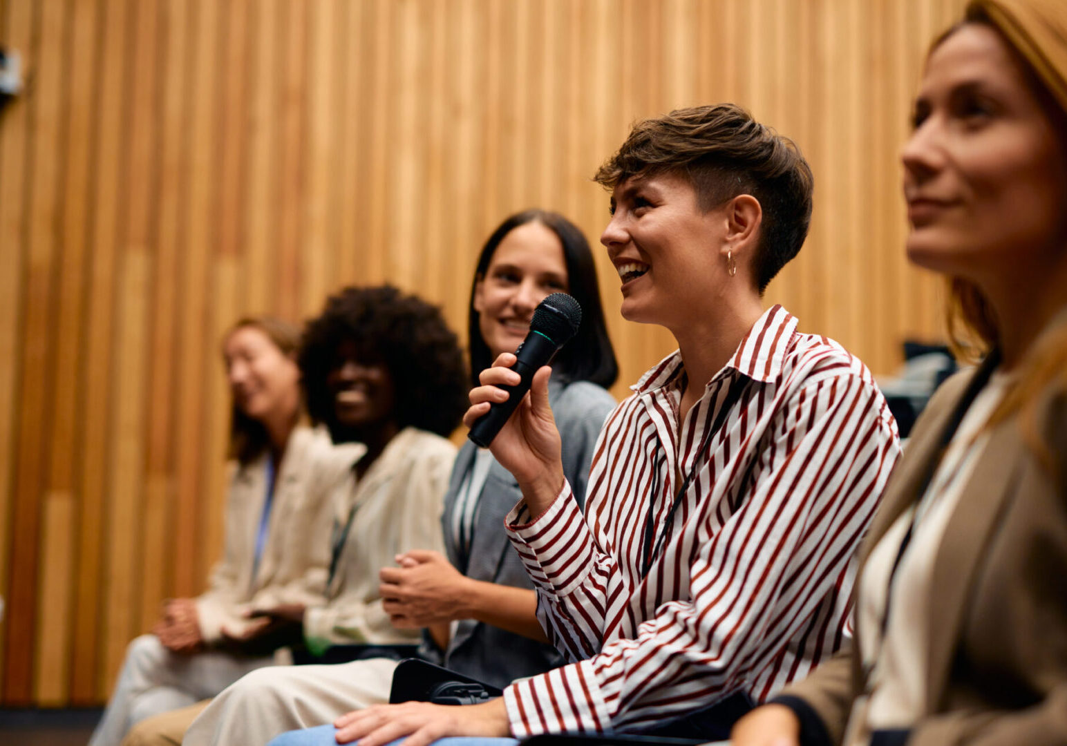 Businesswoman holding a microphone and speaking at a conference, engaging with a group of fellow businesswomen in a modern conference room, fostering discussion and collaboration