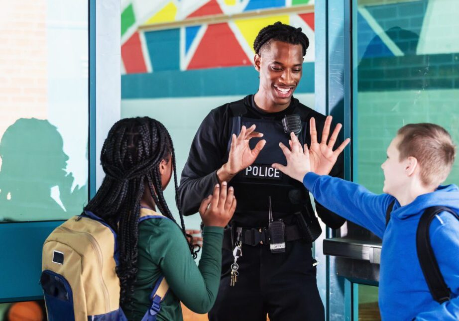 A school resource officer greets students arriving with their backpacks. They are standing face to face with the view over students' shoulders. The officer, a young African-American man, is giving a high-fives to the 10 year old girl and 12 year old boy.